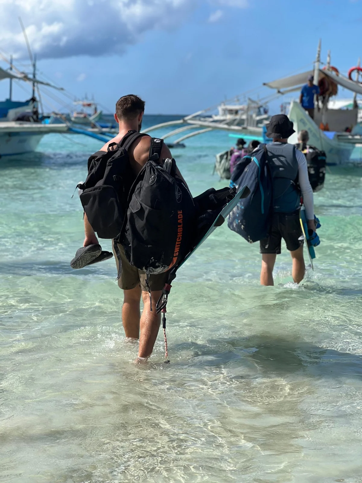 Riders carrying boards and equipment through shallow water toward a boat