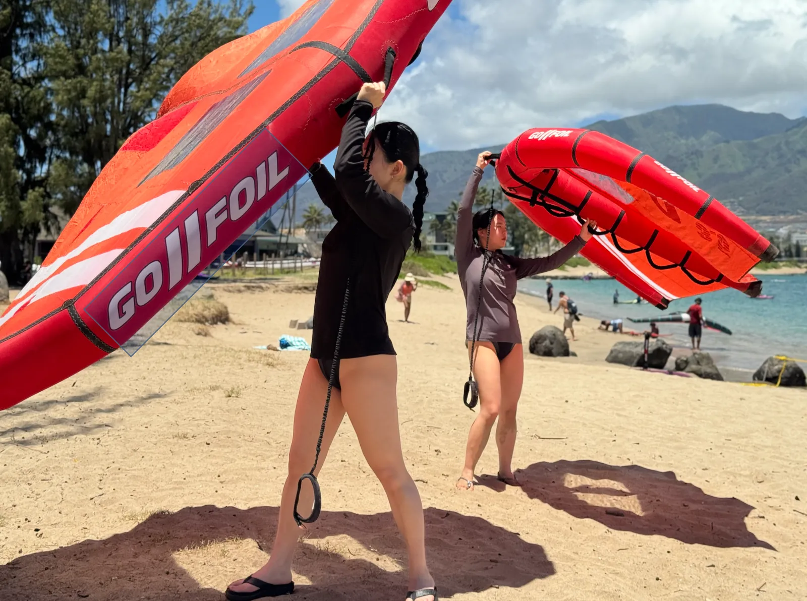 Two riders practicing wing handling on a Maui beach with mountains behind